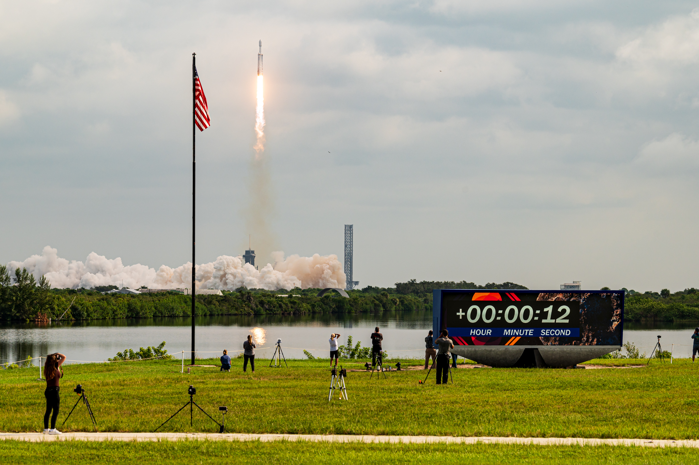 A wide shot of Falcon Heavy and Psyche lifting off. The countdown clock and American flag are in view, along with some other members of the media.