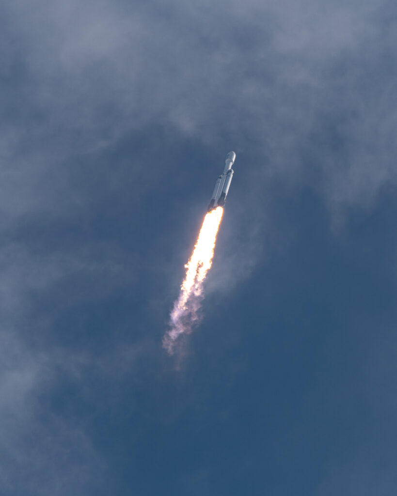 A Falcon heavy rocket ascends into the sky behind three columns of flames.