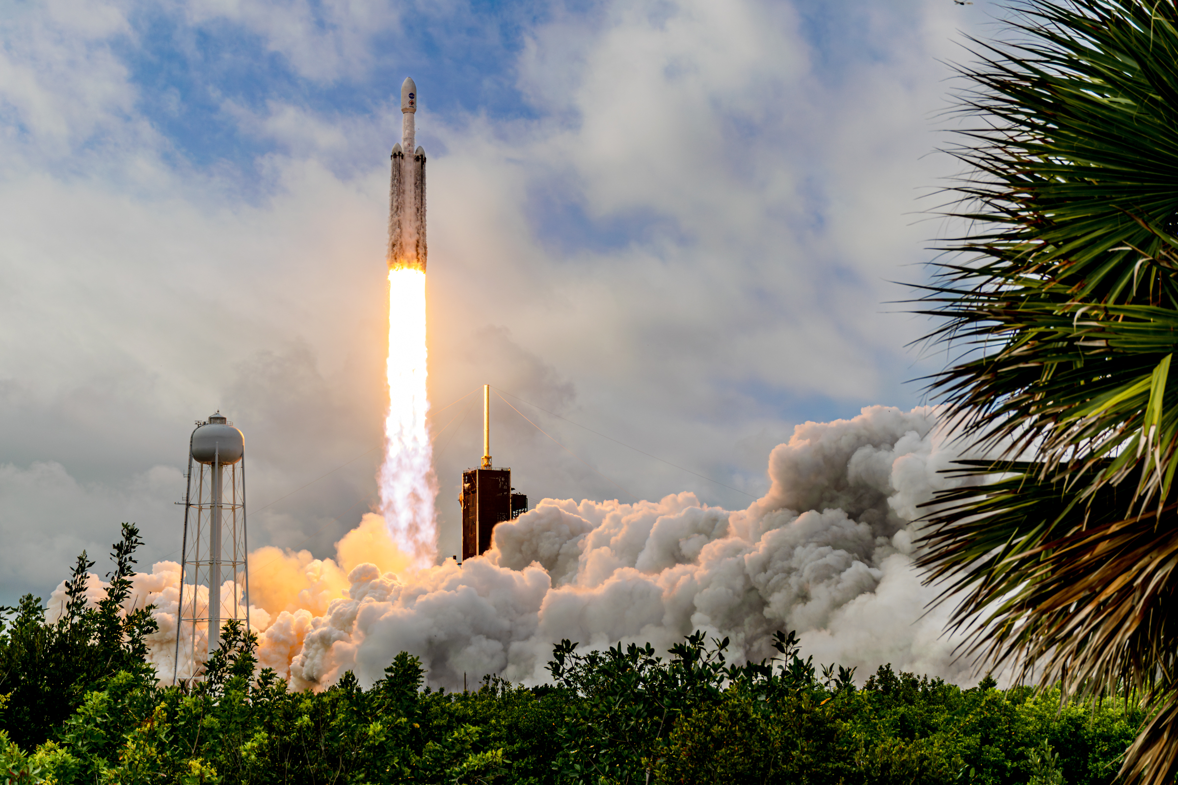 A Falcon Heavy rocket lifts off from Launch Complex-39A at NASA's Kennedy Space Center. There is mangroves and a palm tree in the foreground. The rocket is just clearing the tower and the exhaust plume is expanding towards the camera.